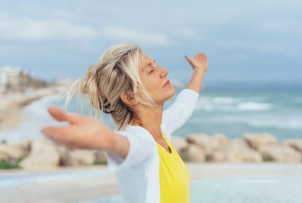 This is a stock photo of a woman on a beach with her arms outspread and face to the sky. It accompanies a news item on Hyatt Hotels launch of its Wellbeing Collective (October 2023). Photo is by stockfour | Canva.