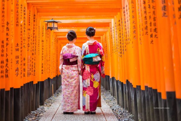This is an image of two geishas walking through red wooden Torii Gate at Fushimi Inari Shrine in Kyoto, Japan. Photo by kasto | Canva.