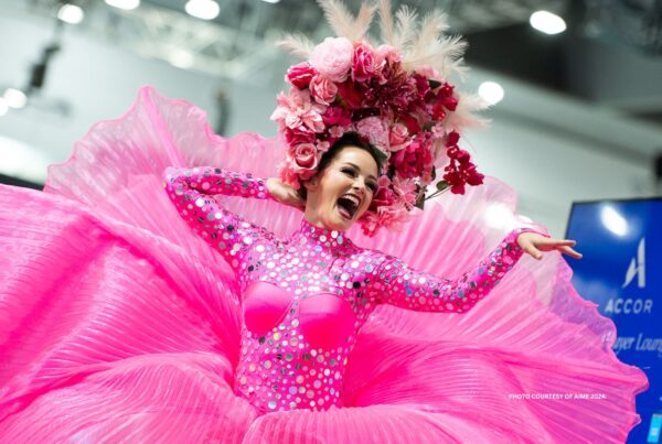 This is an image of a stilt walker in a bright pink costume with floral head dress, performing in the aisles of AIME 2024 (Melbourne, Australia). Photo courtesy of AIME.