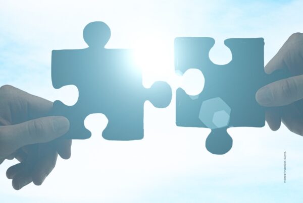 This is a stock image showing hands holding puzzle pieces against a blue sky/bright light background. Theme is partnership. Photo by peshkova | Canva.