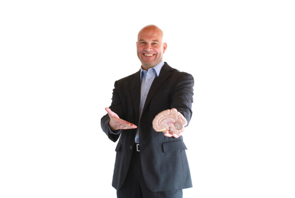 Neuromarketing and events is the subject of one session at the 2022 edition of IBTM World, running November 29-December 1 in Barcelona, Spain. This is an image of the session speaker, Dr. Thomas Trautman. He is dressed in a grey suit and standing with his hand extended and holding a model of a human brain. Photo courtesy of IBTM World.