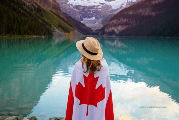 Berkshire Hathaway Travel Protection safest destinations report places Canada at the top of the country rankings. This is a stock image of a woman looking at a lake in Banff, Alberta, Canada. The woman is wrapped in a Canadian flag and wears a straw hat. Photo by Andre Furtado | Canva.